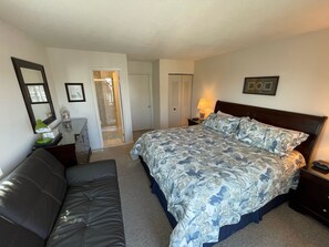 A bedroom with a bed, a sofa, a nightstand, a lamp, a mirror, and a door leading to a bathroom.