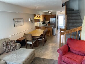 A spacious living area with a dining table, wooden chairs, a red armchair, a sofa, and a kitchen in the background.