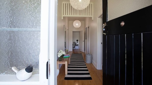 A hallway with a black door, white walls, a wooden floor, a black and white striped rug, a white bench with a vase of flowers, and a hanging light fixture. - THE WHITE HOUSE luxury villa embedded in history (Penola)