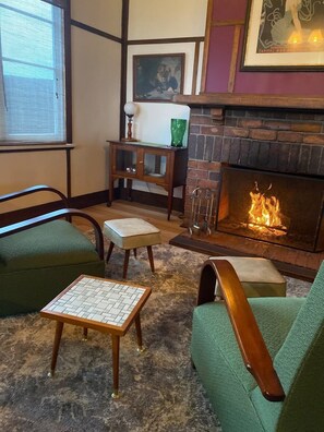 Un salon chaleureux avec une cheminée, des fauteuils à revêtement vert, une petite table et un tableau encadré au mur.