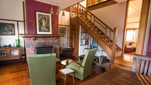 Ein gemütliches Wohnzimmer mit Kamin, grünen Sesseln, einer Holztreppe und einem Bücherregal.