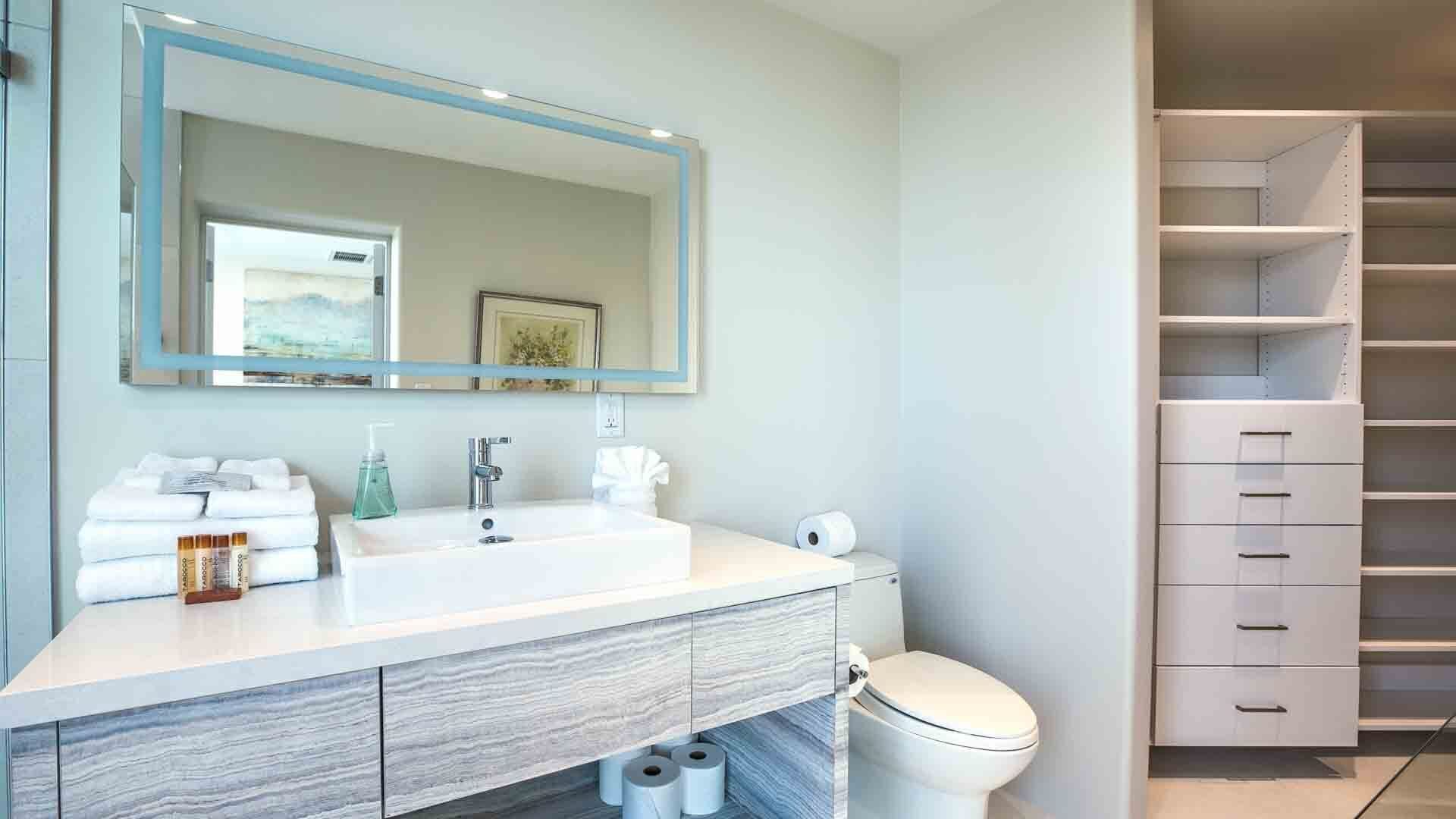 Une salle de bain moderne avec un meuble-lavabo blanc, un grand miroir et des toilettes. On y trouve des serviettes et des articles de toilette sur le meuble-lavabo.