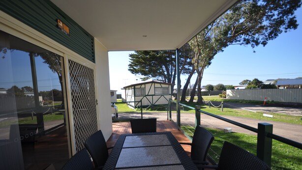 A covered patio with a table and chairs, a view of a building, and a fenced area with trees. - Dylene Caravan Park - 2 Br Family Cabin With Air (Portarlington)
