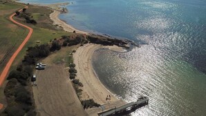 An aerial view of a coastal area with a sandy beach, a dirt road, and a pier extending into the water. - Dylene Caravan Park - 2 Br Family Cabin With Air (Portarlington)
