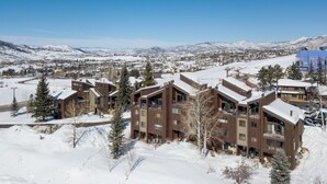 A modern ski resort with wooden buildings, snow-covered surroundings, and a clear view of the mountains in the background. - Walk to Gondolas! Heated Pool & Hot Tubs - Perfect for 2 couples & 2-4 singles or kids (Steamboat Springs)