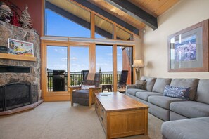 A cozy living room with a fireplace, large windows, a wooden coffee table, and a gray sofa.