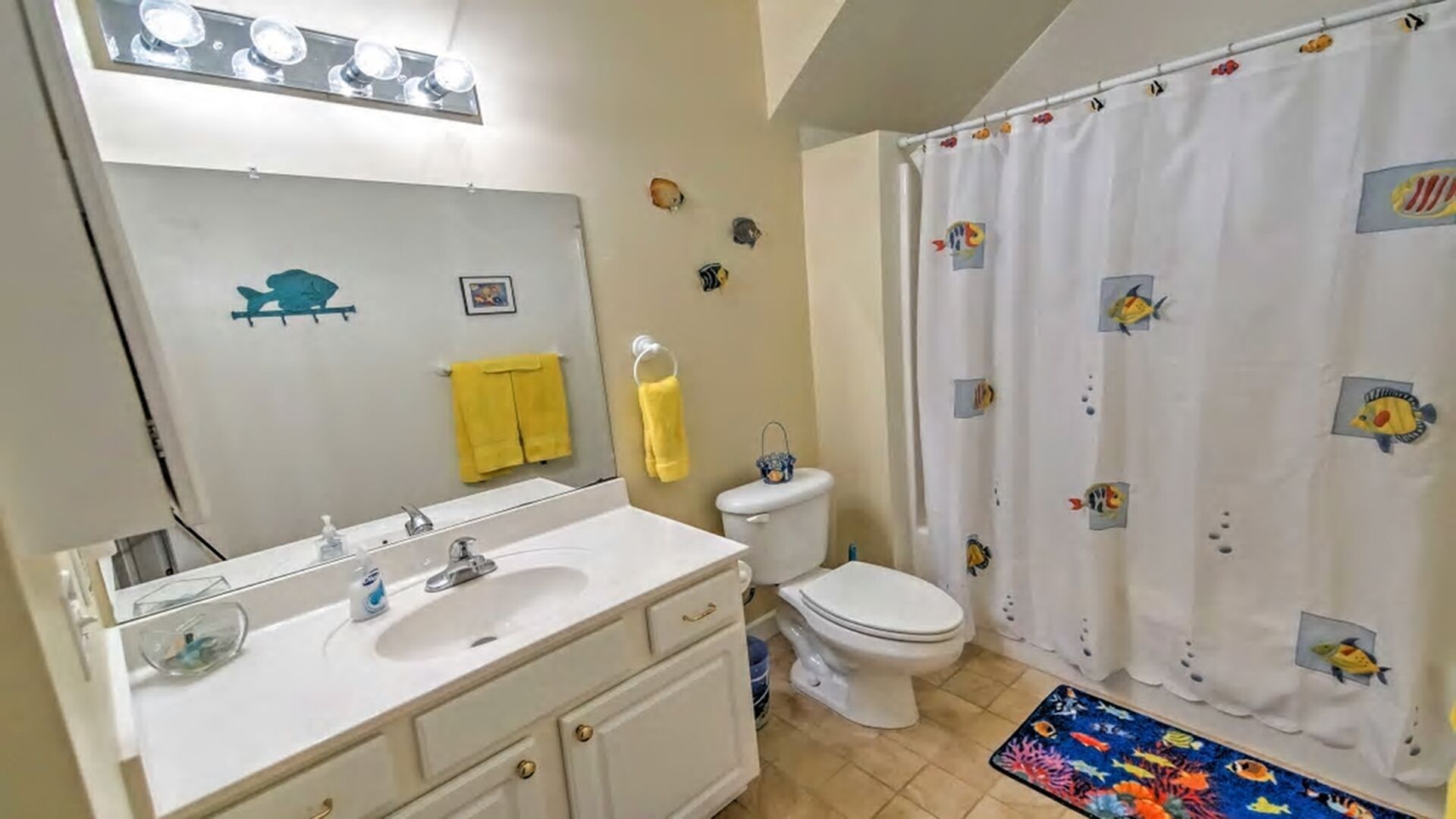 Un baño con lavamanos blanco, espejo e inodoro. Toallas amarillas cuelgan de la pared y una cortina de ducha con diseños de peces.