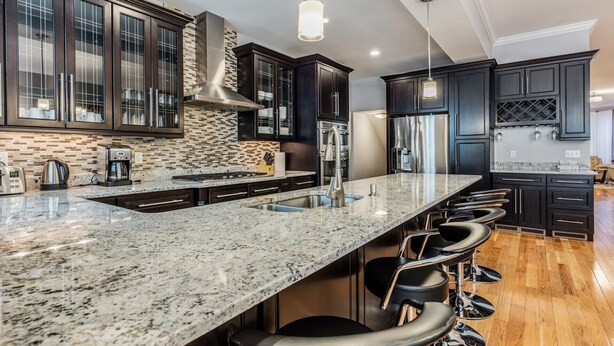 A modern kitchen with dark cabinetry, a large island with bar stools, stainless steel appliances, and a patterned backsplash. - UltraLuxe Apt in Boston Brownstone in Prime Area (Brookline)
