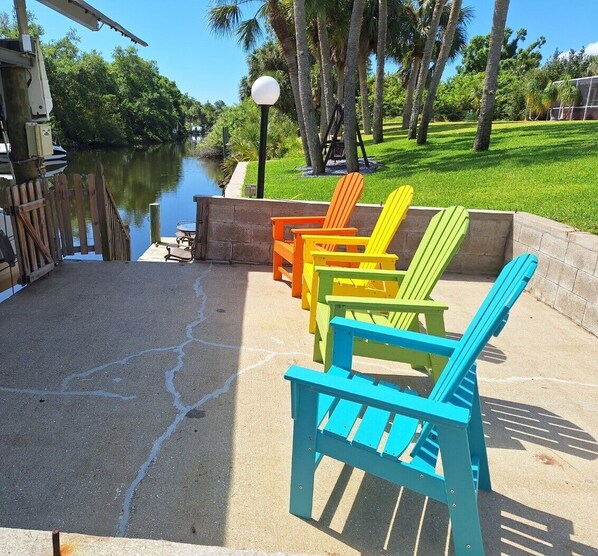 A patio area with colorful Adirondack chairs, a concrete floor, a wooden fence, and a body of water in the background.