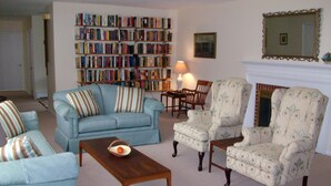 Een woonkamer met een bank, fauteuils, een salontafel, een open haard en een boekenplank.