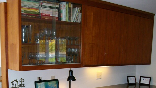 Een kleine keuken met houten kastjes, een laptop op het aanrecht en een stoel.