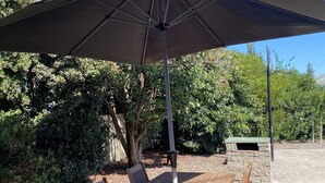 A wooden table and chairs under a large umbrella, set on a paved patio with stone walls and greenery in the background. - Torquay Shores - Family, Friends, Pets Welcome! (Torquay Surfcoast)