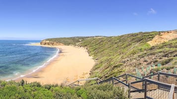 A coastal landscape with a sandy beach, a wooden staircase leading down to the beach, and a clear blue sky.