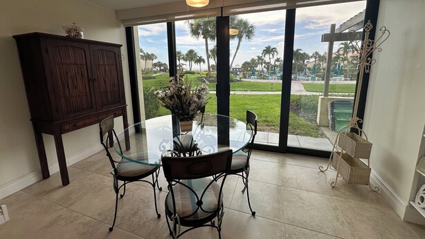 A dining area with a glass table and chairs, a dark wooden cabinet, and a view of a landscaped outdoor area with palm trees and a pathway. - New Listing! Ground floor, easy access to the pool and beach! Siesta Dunes 2-104 (Siesta Key)