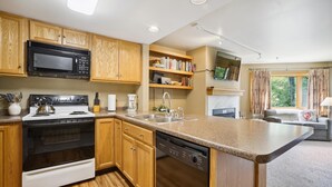 Cozinha moderna com armários de madeira, micro-ondas, fogão e lava-louças. Há também uma máquina de café, pia e uma TV fixada na parede.