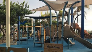 Een speelplaats met een blauwe, rubberen ondergrond, een glijbaan en een bordje waarop staat dat het gebied bedoeld is voor kinderen van 2 tot 5 jaar, met begeleiding van een volwassene.