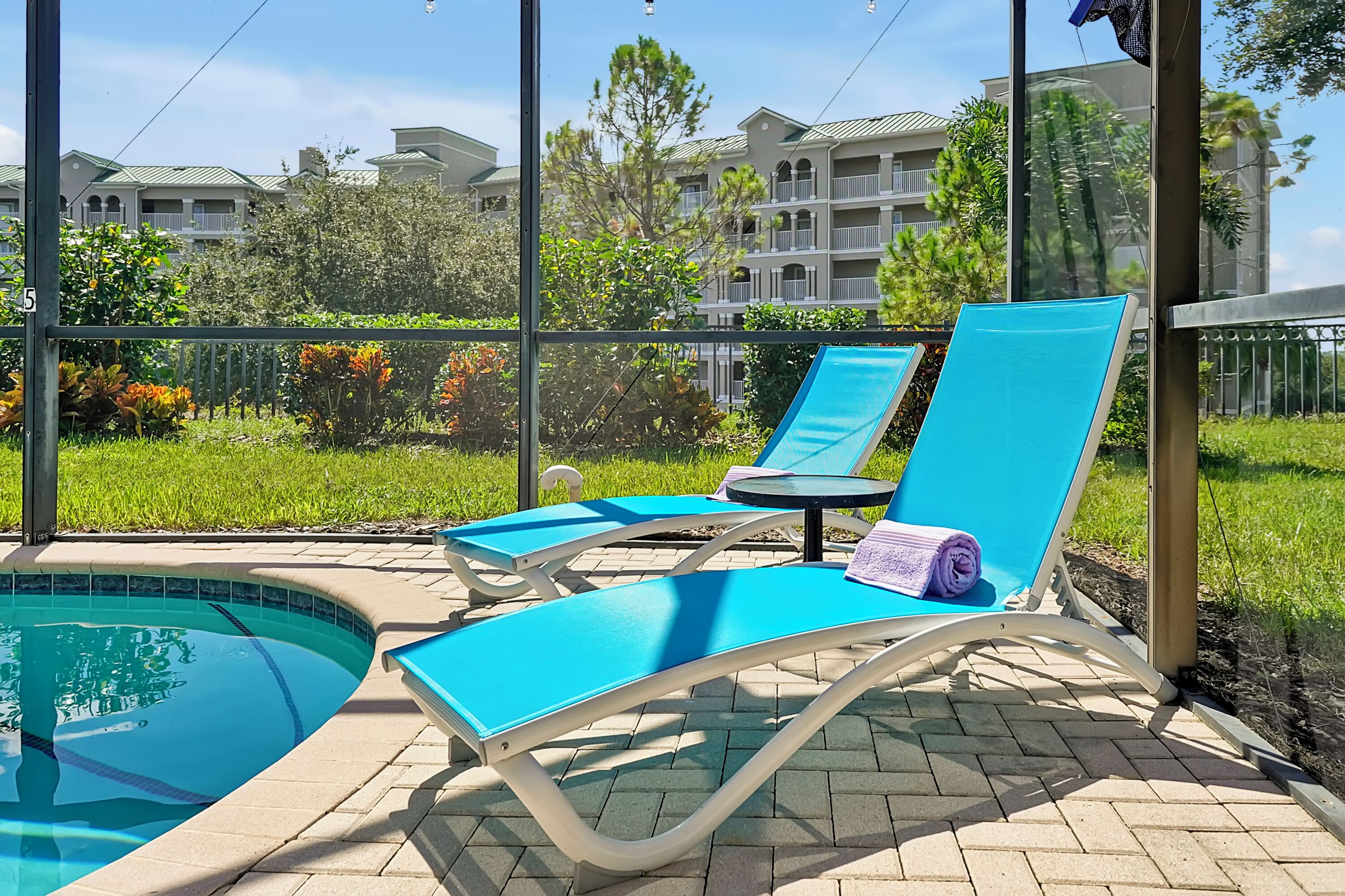 Un espace aménagé au bord de la piscine, avec deux chaises longues bleues, dont l’une est recouverte d’une serviette, et d’où l’on a une vue dégagée sur un immeuble d’habitation en arrière-plan.