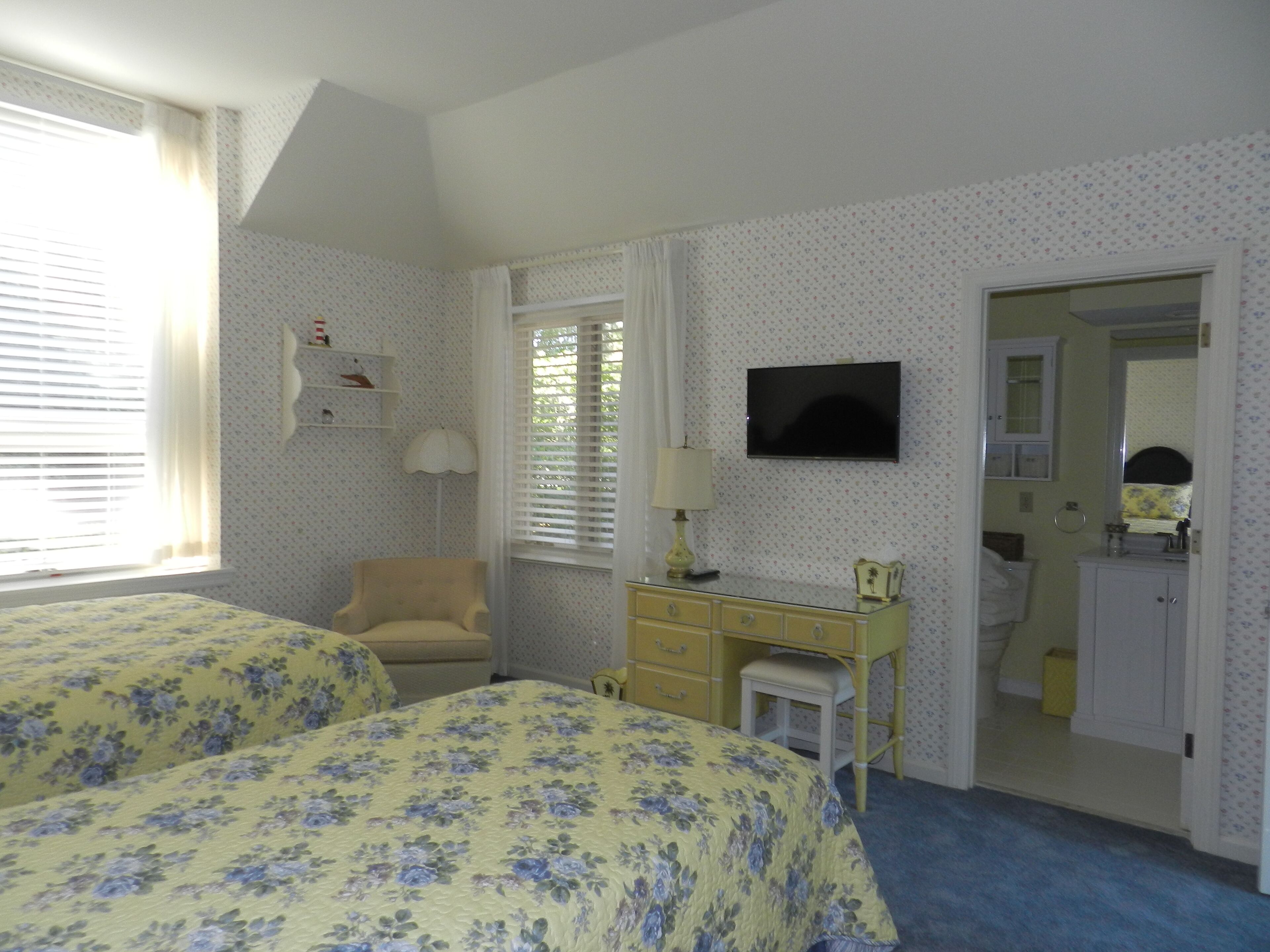 A bedroom with two beds, a desk, a chair, a television, and a bathroom visible through an open door.