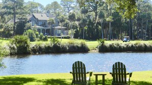 A serene lakeside setting with two Adirondack chairs facing a calm water body, surrounded by lush greenery and a distant golf course. - Rates Jan through mid March 2027 are reduced for longer stays. (Hilton Head Island)