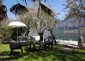 An outdoor seating area with a table and chairs, a white umbrella, and a view of a lake with mountains in the background.