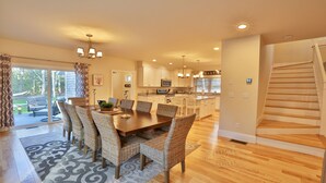A spacious dining area with a large wooden table and wicker chairs, a kitchen with white cabinets, and a staircase leading to another level. - Modern, luxury 5-bed, 4.5-bath w/full basement! Now booking for 2026! (Oak Bluffs)