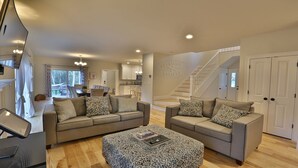 A spacious living room with two sofas and a patterned ottoman, a staircase, and a kitchen in the background. - Modern, luxury 5-bed, 4.5-bath w/full basement! Now booking for 2026! (Oak Bluffs)
