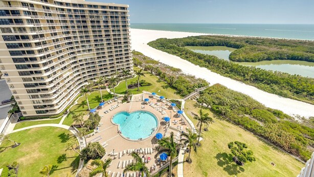 A hotel with a large pool surrounded by lounge chairs and umbrellas, a beach, and a mangrove area.