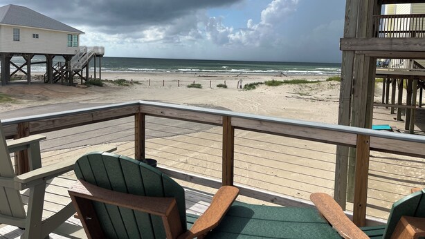 A wooden deck with chairs overlooking a beach, a house on stilts, and a cloudy sky.