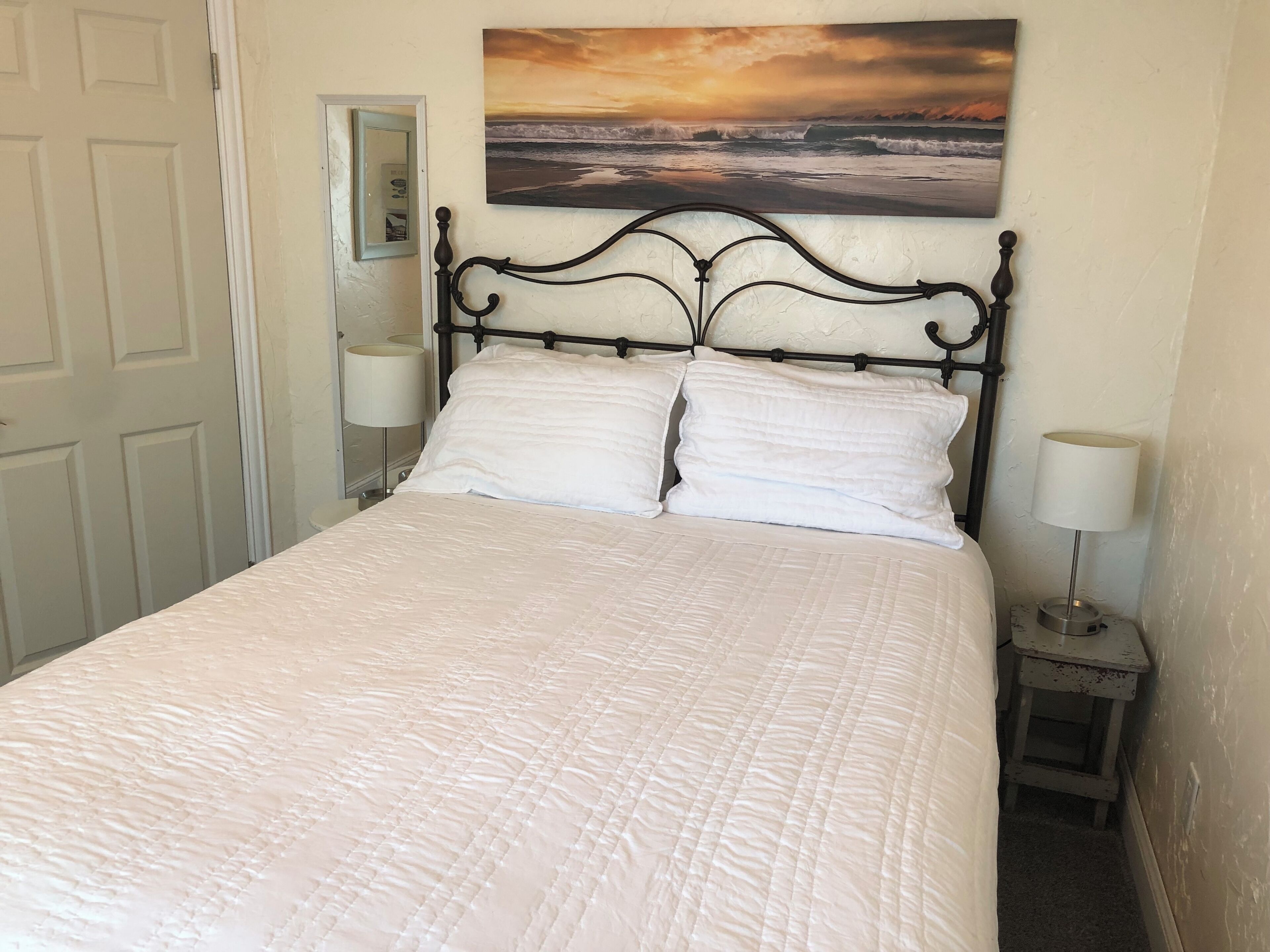 Un lit bien fait, avec une couette blanche matelassée, une tête de lit en métal noir et, au mur derrière, un tableau représentant un coucher de soleil sur l’océan.