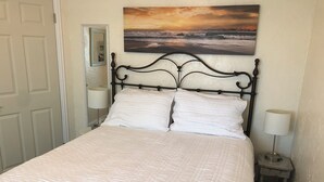 A neatly made bed with a quilted white duvet, a black metal headboard, and a painting of a sunset over the ocean on the wall behind it.