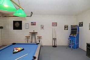 Une table de billard avec des billes, un jeu de fléchettes et un juke-box dans une pièce aux murs blancs et au sol bleu.