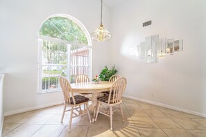 A dining area with a round table, four chairs, a hanging light fixture, and a large window with a view of greenery.
