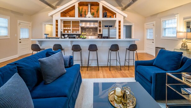 Ein geräumiges Loft mit hoher Decke, freiliegenden Holzbalken, einer zentral gelegenen Küche mit Bar und einer gemütlichen Sitzecke mit blauen Sofas.