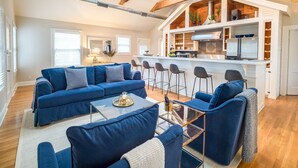 Ein geräumiges Wohnzimmer mit blauem Sofagarnitur, einem Glastisch und einer Küchenzeile mit Barhocker.