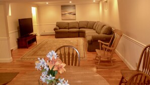 A living room with a sectional sofa, a flat-screen TV, wooden chairs, and a dining table with a floral centerpiece. - Walk to the Beach, Heated Salt Water Pool, Central AC (Dennis)