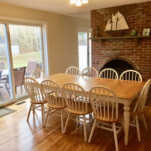 A dining area with a wooden table and white chairs, a brick fireplace, and a view through a glass door to the outdoors.