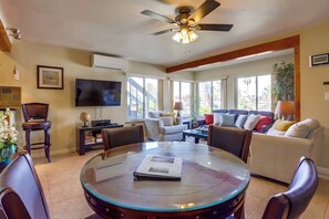 A spacious living room with a round glass table, comfortable seating, a ceiling fan, and large windows.