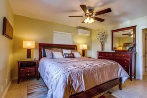 A bedroom with a bed, bedside tables, a ceiling fan, an air conditioning unit, a mirror, and a vase with flowers.
