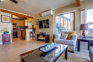 A modern living room with a glass coffee table, a sofa with cushions, a television, and a kitchen area with wooden cabinets and a stove.