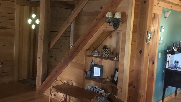 A cozy cabin interior with a wooden staircase, a small table with chairs, a shelf with decorative items, and a desk with a computer.