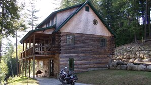 A log cabin with a balcony, a stone retaining wall, and a motorcycle parked in front.