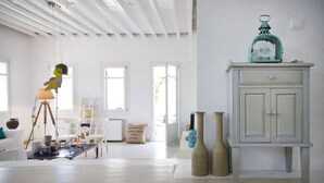 Um quarto claro e espaçoso, com teto branco, piso de madeira e um armário branco com um candeeiro verde-água em cima. Há dois vasos no chão e um abajur com uma escultura de pássaro sobre um tripé.