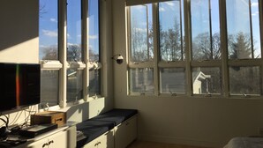A bedroom with large windows, a bed, a desk with a computer, and a TV.