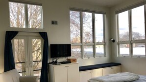A bedroom with a bed, a TV, a desk, and a window with a view of trees and a building.