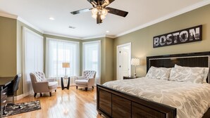 Une chambre avec un grand lit, deux fauteuils, une petite table et un ventilateur de plafond. Un panneau avec l’inscription « Boston » est accroché au mur.