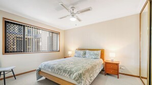 Ein Schlafzimmer mit Bett, Nachttischlampen, einem Nachttisch, einem Sessel, einem Fenster mit Gitterstäben und einem Deckenventilator.