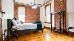 A bedroom with a bed, a standing lamp, a ceiling fan, and a brick wall.