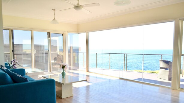 A spacious living room with a large sliding glass door leading to a balcony with a view of the ocean. - Kiama Dream – Absolute Waterfront | Panoramic Ocean Views | Large Groups (Kiama)