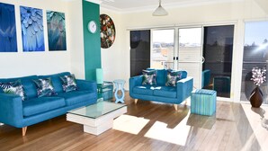 A modern living room with two blue sofas, a glass coffee table, and a view through large sliding glass doors to an outdoor balcony. - Kiama Dream – Absolute Waterfront | Panoramic Ocean Views | Large Groups (Kiama)