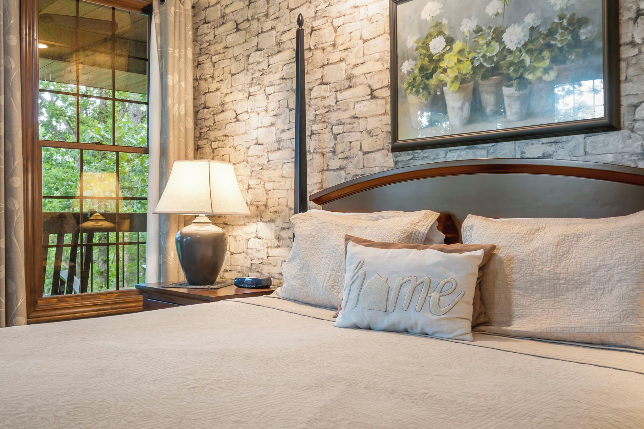 A bedroom with a stone wall, a bed with a headboard, a nightstand with a lamp, and a window with a view of greenery.